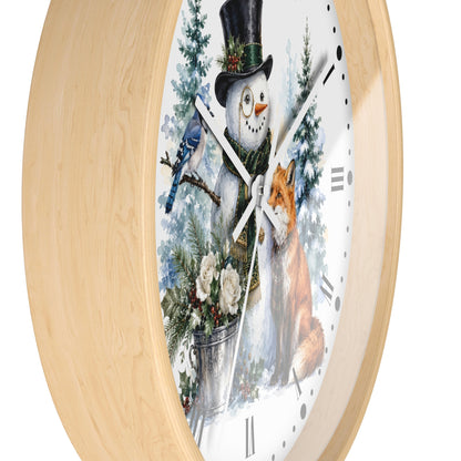Round wall clock with a light natural wood frame and a white face featuring a winter illustration of a snowman in a top hat, a bluebird, a fox, evergreen trees, and a bucket of flowers.