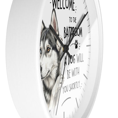 A white round wall clock with a husky dog illustration and the humorous text 'WELCOME TO THE BATHROOM A DOG WILL BE WITH YOU SHORTLY' printed on the face.