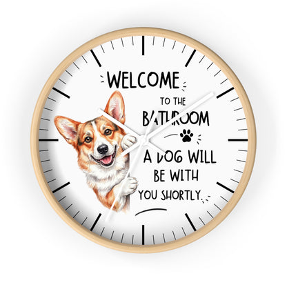 A round wall clock with a light wood frame and a white face, featuring a cartoon Corgi illustration and the text 'WELCOME TO THE BATHROOM A DOG WILL BE WITH YOU SHORTLY.' on a plain white background.