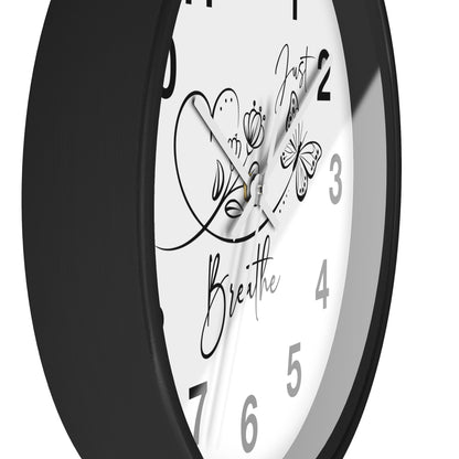 A close-up of a round wall clock with a black frame and a white face featuring black Arabic numerals and decorative script text reading 'Just Breathe' with butterfly and heart illustrations.