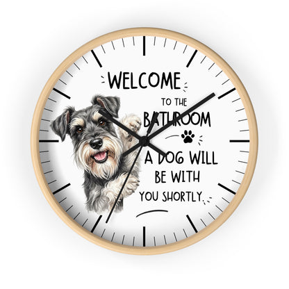 A round wall clock with a light wood frame and a white face, featuring a sketch of a gray and white schnauzer dog and the text 'WELCOME TO THE BATHROOM A DOG WILL BE WITH YOU SHORTLY.' on a plain white background.