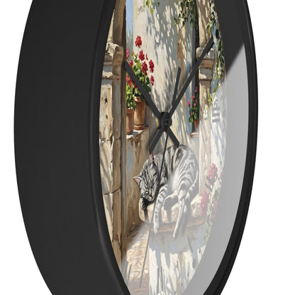 Round wall clock with a black frame and a printed illustration of a gray tabby cat lounging on a stone ledge in a sunlit courtyard, with red flowers and green vines around it.
