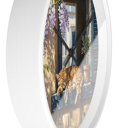 An oval wall clock with a white frame and black hands, featuring a printed illustration of an orange tabby cat sleeping on a windowsill with purple wisteria flowers and a sunlit Mediterranean-style courtyard.