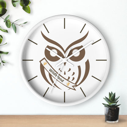A round wall clock with a white face and white frame, featuring a brown owl illustration holding a banner that reads 'Wild Food'. The clock is mounted on a white wall above a light wood surface, with green leaves and a small succulent plant as decorative props.