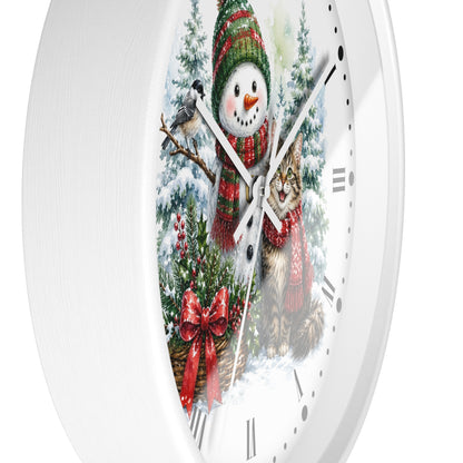 A white round wall clock with a festive winter illustration on the face, showing a snowman in a green and red knit hat and scarf beside a tabby cat in a red scarf, with snowy trees, a bird, and a large red bow. Roman numerals are visible on the clock face.