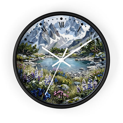 A round wall clock with a black frame and a scenic mountain landscape printed on the face. The artwork shows snow-capped blue-gray mountains, a lake, green trees, and wildflowers in the foreground.