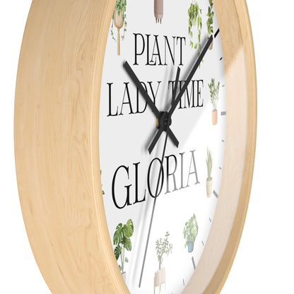 Round wall clock with a light natural wood frame, a white face, black hands, and the phrase "PLANT LADY TIME GLORIA" surrounded by small potted plant illustrations.