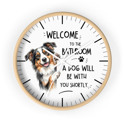 A round wall clock with a light wood frame and a white face, featuring a printed illustration of a brown, white, and black dog next to the text 'WELCOME TO THE BATHROOM A DOG WILL BE WITH YOU SHORTLY.' The clock is shown against a plain white background.