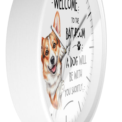 A white round wall clock with a printed illustration of a corgi dog and the text 'WELCOME TO THE BATHROOM A DOG WILL BE WITH YOU SHORTLY' on the front.