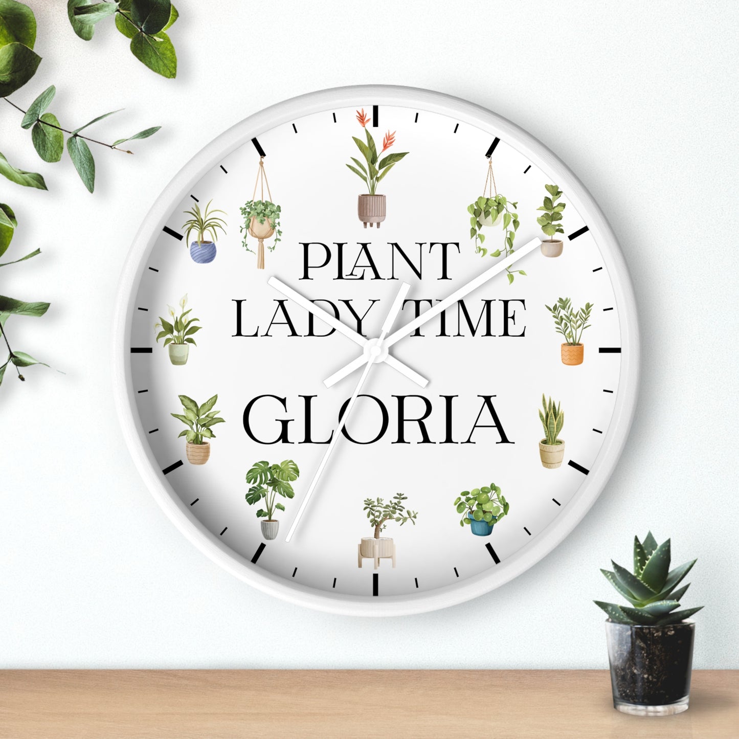 A round white wall clock with black hands and black hour markers, featuring the text 'PLANT LADY TIME GLORIA' and illustrated hanging plants around the face. The clock is shown hanging on a white wall above a wooden surface, with decorative green leaves and a small potted succulent nearby.