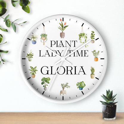 A round white wall clock with black hands and black hour markers, featuring the text 'PLANT LADY TIME GLORIA' and illustrated hanging plants around the face. The clock is shown hanging on a white wall above a wooden surface, with decorative green leaves and a small potted succulent nearby.
