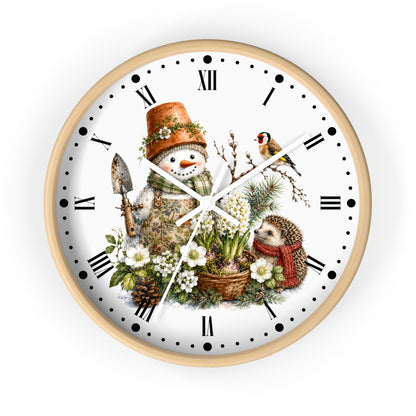A round wall clock with a light wood frame and a white face featuring a whimsical winter garden illustration. The design includes a snowman in a terracotta pot hat, a small bird, a hedgehog in a red scarf, white flowers, pinecones, and Roman numerals with black dot hour markers.
