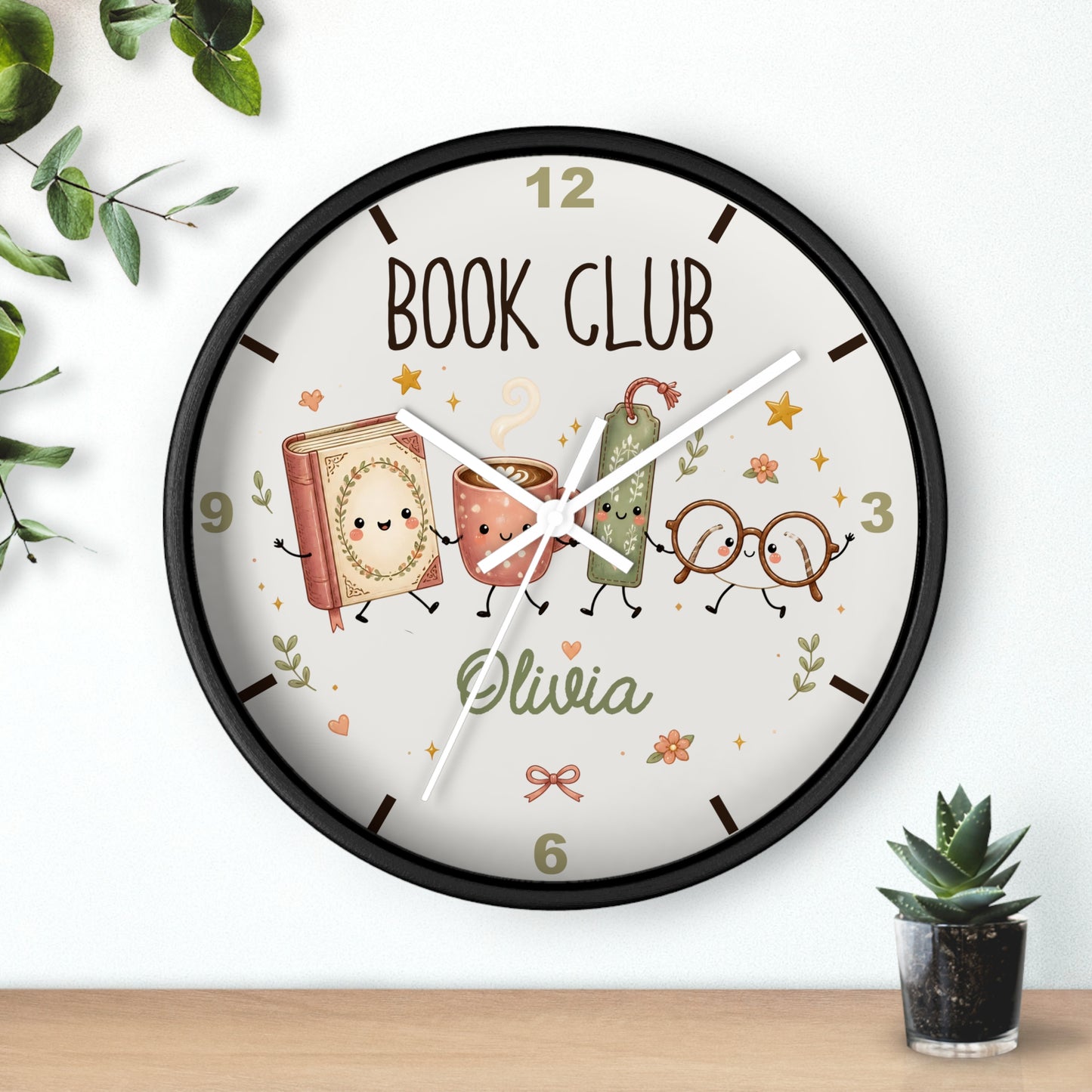 A personalized round wall clock with a black frame, featuring a cream-colored face with 'BOOK CLUB' text, cartoon book and cup characters, glasses, and the name 'Olivia'. The clock is shown hanging on a white wall above a wooden shelf with small plants.