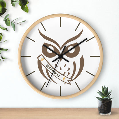 A round wall clock with a light wood frame and a white face featuring a large brown owl graphic and the words 'Wildwood' on a banner. The clock is mounted on a white wall above a light wood surface, with decorative green leaves on the left and a small potted succulent on the right.
