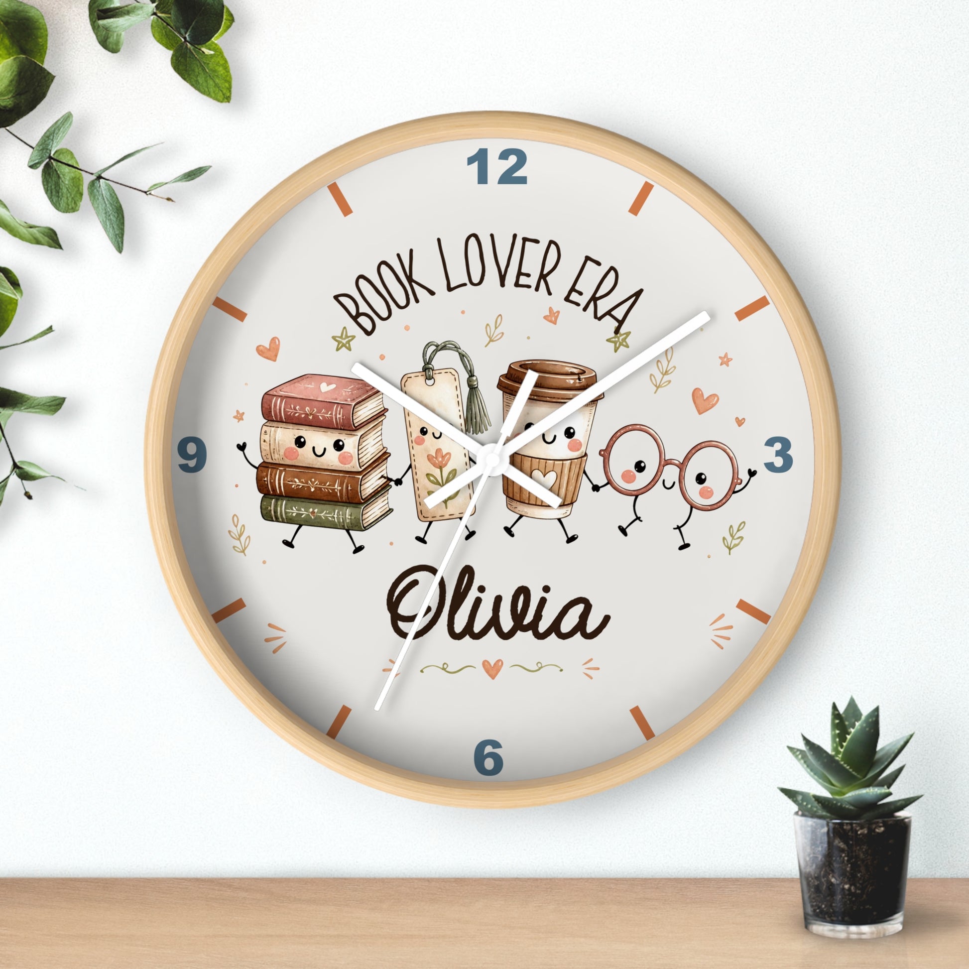 A round wall clock with a light wood frame and a white face, featuring cartoon book and coffee cup characters and the text 'BOOK LOVER ERA' and the name 'Olivia'. The clock is mounted on a white wall above a wooden surface, with small green plants in the corners.