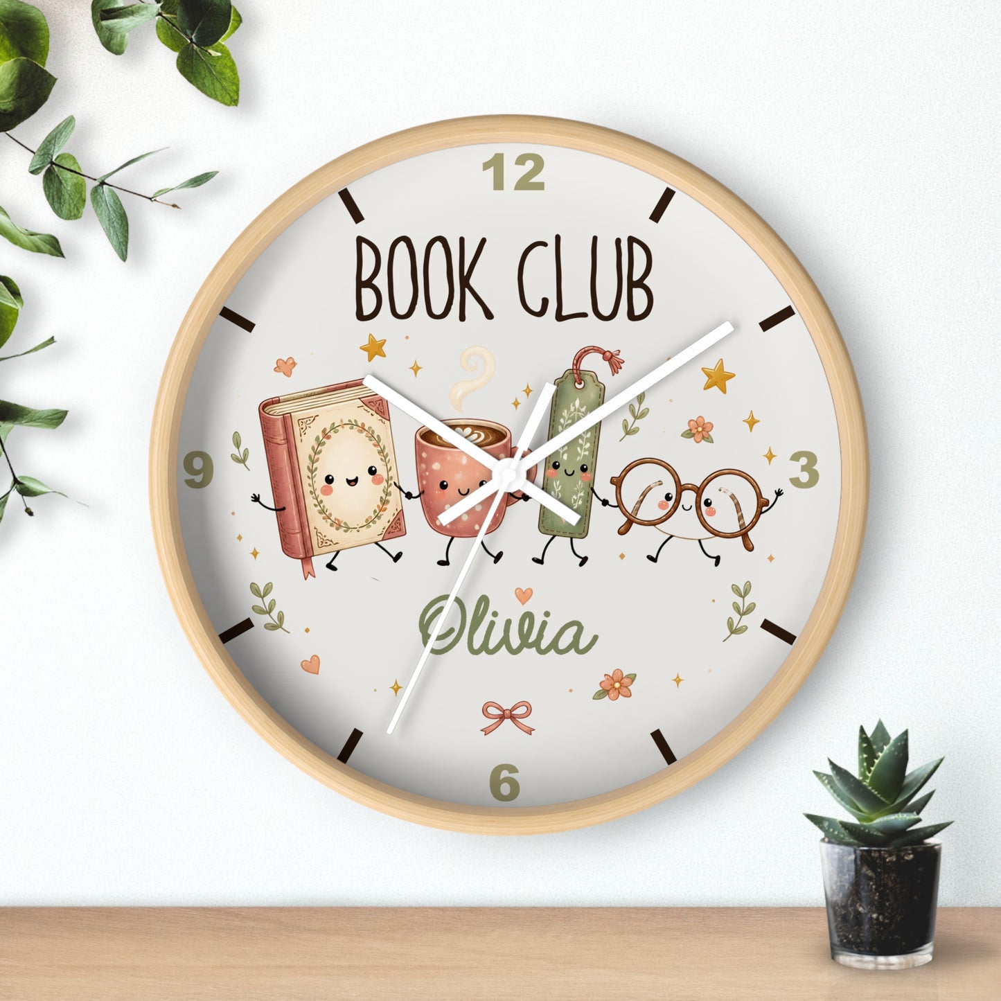 A round wall clock with a light wood frame and a cream-colored face, featuring a 'BOOK CLUB' design with cute illustrated characters and the name 'Olivia'. The clock is shown hanging on a white wall above a wooden shelf, with decorative green leaves and a small potted succulent nearby.