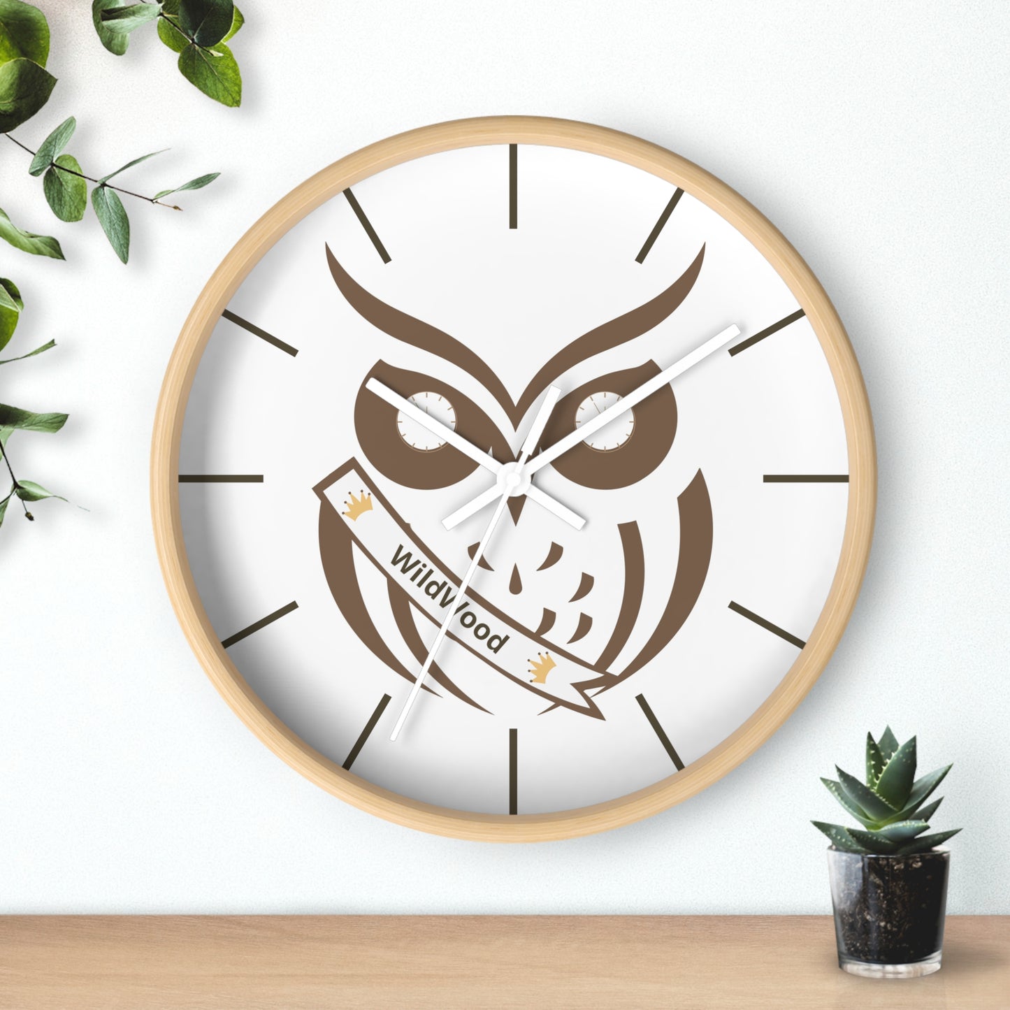 A round wall clock with a light wood frame and a white face featuring a large brown owl illustration and the words 'Wild Good' on a banner. The clock is mounted on a white wall above a light wood surface, with decorative green leaves on the left and a small potted succulent on the right.