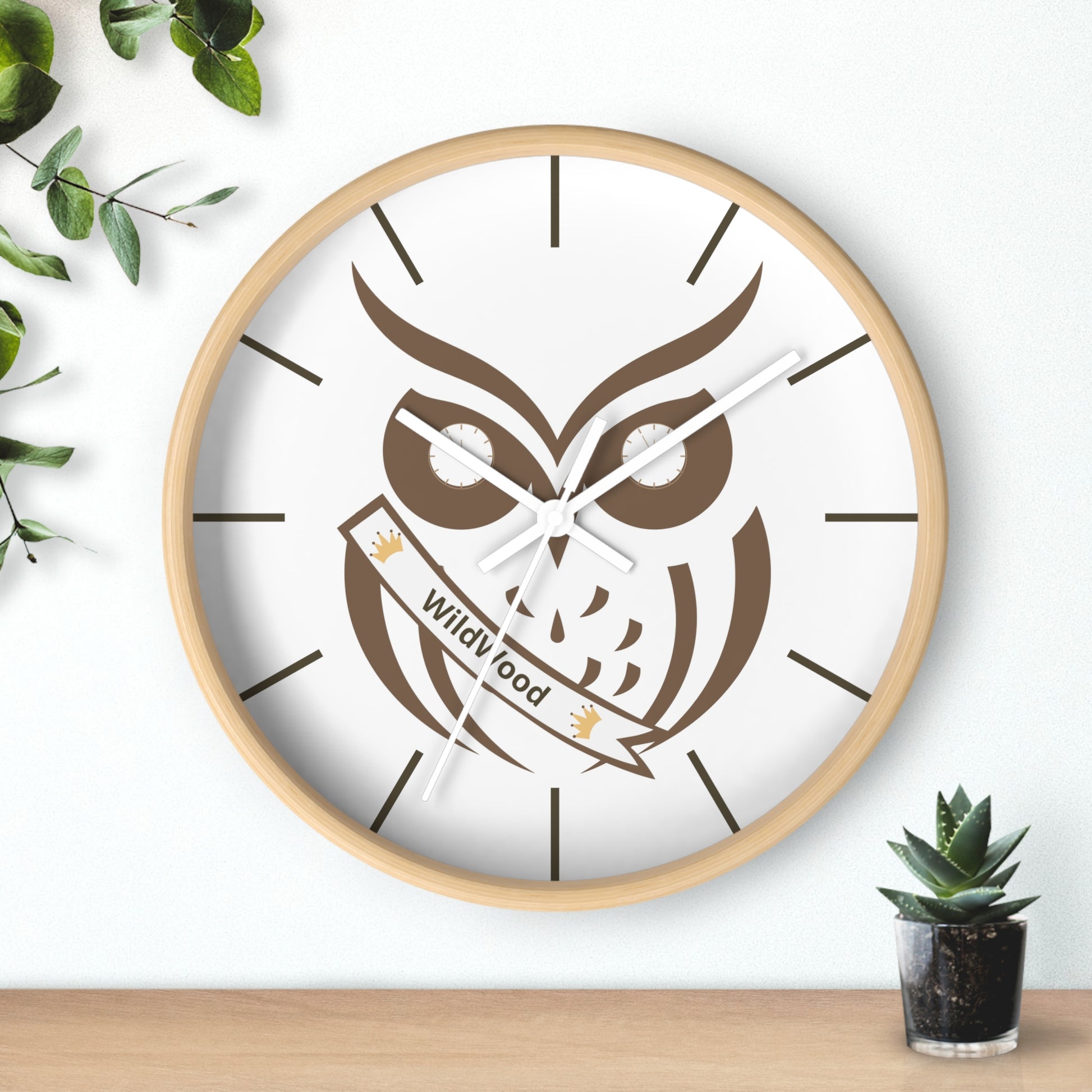 A round wall clock with a light wood frame and a white face featuring a large brown owl illustration and the words 'Wild Good' on a banner. The clock is mounted on a white wall above a light wood surface, with decorative green leaves on the left and a small potted succulent on the right.