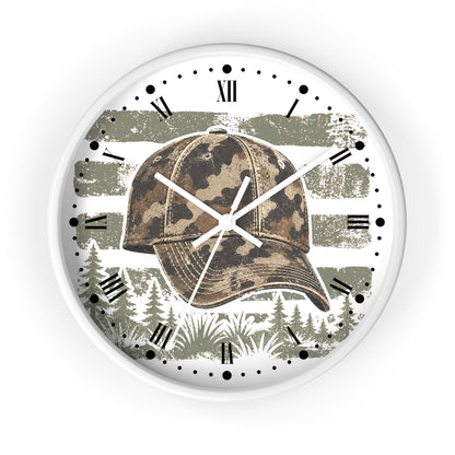 A round wall clock with a white frame and face, featuring a camouflage baseball cap graphic over a distressed olive green and white American flag, with pine trees along the bottom and black Roman numerals around the dial.