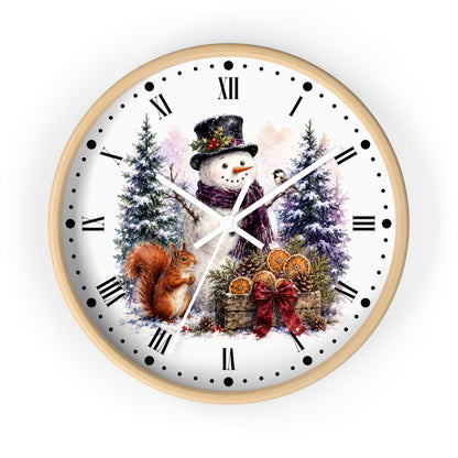 A round wall clock with a light wood frame and a white face featuring a winter holiday illustration of a snowman, a squirrel, evergreen trees, and gift baskets. The clock has black Roman numerals and black dot hour markers with white hands.