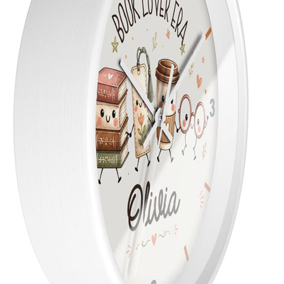 A round white wall clock with a cute cartoon book-themed design. The clock face features an illustration of smiling books, a coffee cup, and glasses, with the text 'BOOK LOVER ERA' at the top and the name 'Olivia' written in a cursive font near the bottom.