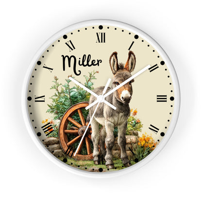 A round wall clock with a white frame and a cream-colored face featuring a detailed illustration of a donkey, a wooden wagon wheel, greenery, and the name 'Miller' in black script. The clock has black Roman numerals and white hands, shown against a plain white background.