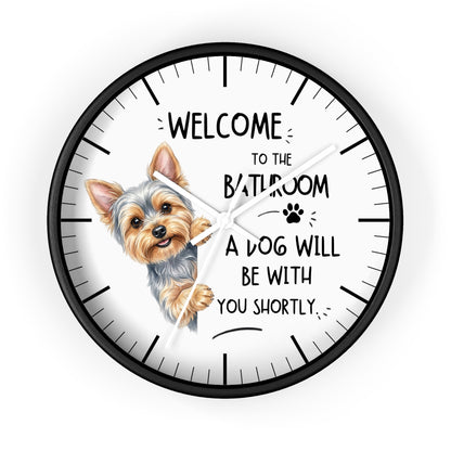 A round wall clock with a black frame and white face, featuring a small dog illustration and the text 'WELCOME TO THE BATHROOM A DOG WILL BE WITH YOU SHORTLY.' on a light gray background.