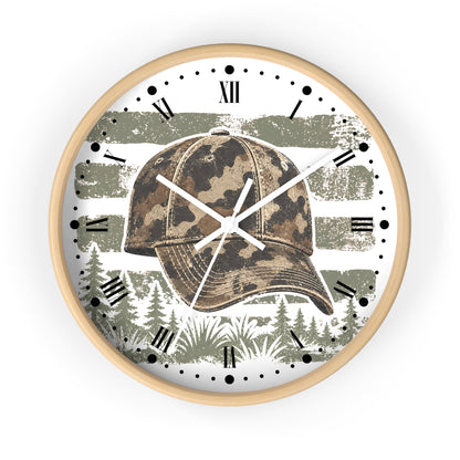 A round wall clock with a light wood frame, featuring a camouflage baseball cap graphic over a distressed gray and white striped background with forest silhouettes and black Roman numerals.