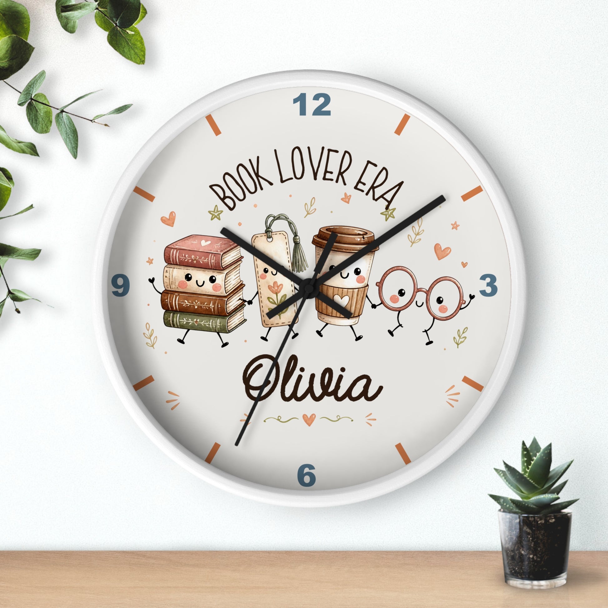A round personalized wall clock with a white frame, featuring cute illustrations of books, coffee cups, and reading glasses on a cream face. The clock is personalized with the name 'Olivia' and the text 'BOOK LOVER ERA'.