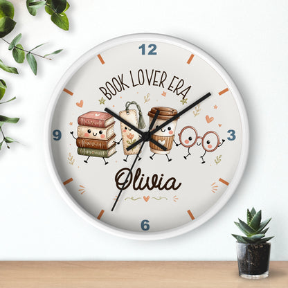 A round personalized wall clock with a white frame, featuring cute illustrations of books, coffee cups, and reading glasses on a cream face. The clock is personalized with the name 'Olivia' and the text 'BOOK LOVER ERA'.
