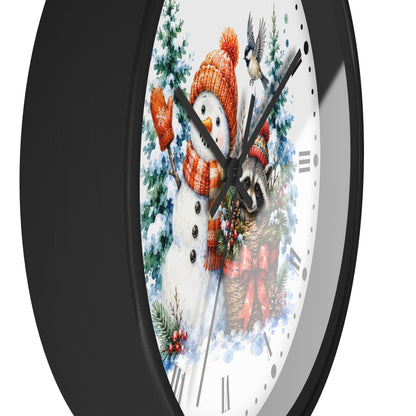 A close-up of a round wall clock with a black frame and a white face featuring a winter holiday illustration of a snowman in an orange knit hat and scarf, evergreen trees, birds, and a red bow.