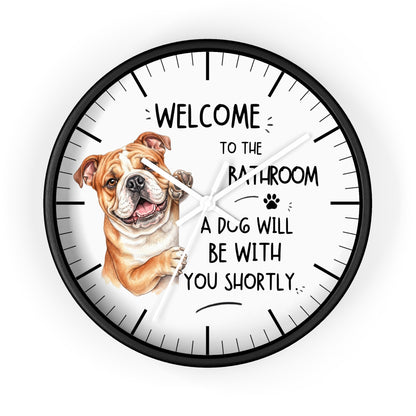 A round wall clock with a black frame and white face, featuring a brown and white bulldog illustration on the left and the text 'WELCOME TO THE BATHROOM A DOG WILL BE WITH YOU SHORTLY.' on a light gray background.