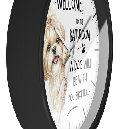 A round wall clock with a black frame and a white face featuring a watercolor illustration of a small fluffy dog and the text 'WELCOME TO THE BATHROOM A DOG WILL BE WITH YOU SHORTLY'.