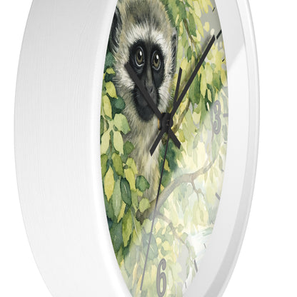 A round white wall clock featuring a watercolor-style print of a vervet monkey surrounded by green leaves, with black hands and gray numerals, shown against a plain white background.