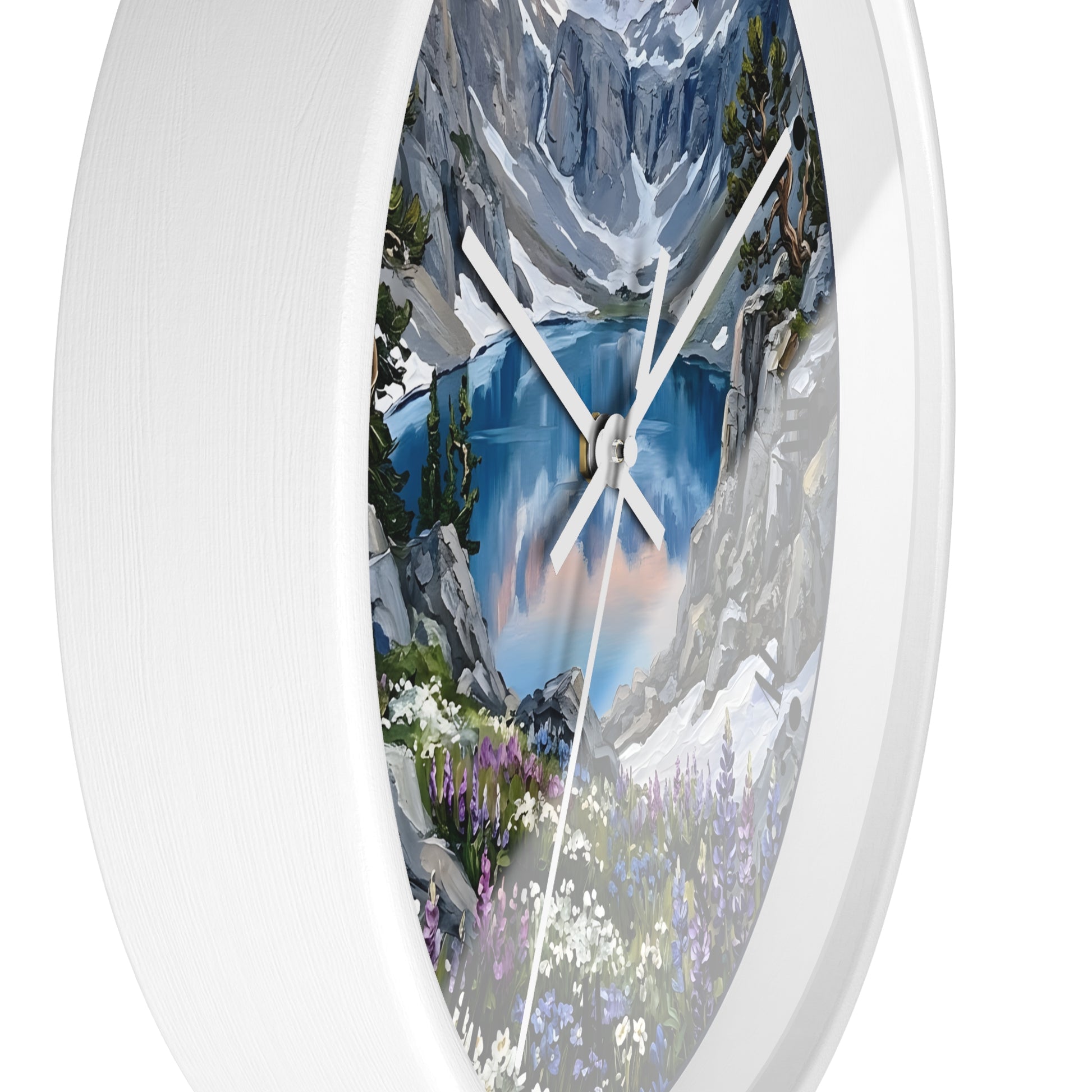 A white round wall clock with a scenic mountain lake print on the face, featuring blue water, gray and white peaks, green trees, and purple wildflowers. The clock is shown against a plain white background.