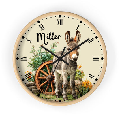 A personalized round wall clock with a light wood frame, featuring a cream-colored face with black Roman numerals and hands. The center of the clock is illustrated with a gray and white donkey standing next to an old wooden wagon wheel, with the name 'Miller' written in black script.