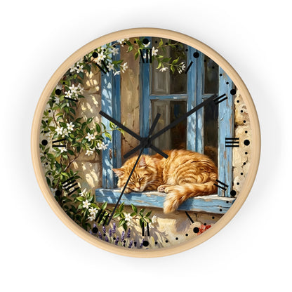 Round wall clock with a light wood frame and a printed illustration of an orange tabby cat sleeping on a blue window sill, surrounded by climbing white flowers and green leaves. The clock face has black Roman numerals and black hands, shown against a plain white background.