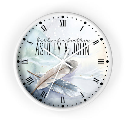 A round wall clock with a white frame and a printed design featuring the text 'Birds of a feather ASHLEY & JOHN' over two large feathers on a soft blue and white watercolor background. The clock face has black Roman numerals and black dot hour markers, with white hands.