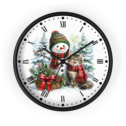 A round wall clock with a black frame and Roman numerals, featuring a festive Christmas illustration on the white clock face. The design shows a snowman in a green and red knit hat and scarf next to a gray tabby cat wearing a red knit scarf, with snowy trees and a basket of holly in the background.