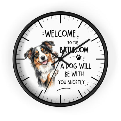 A round wall clock with a black frame and a white face featuring a watercolor illustration of an Australian Shepherd dog and the humorous text 'WELCOME TO THE BATHROOM A DOG WILL BE WITH YOU SHORTLY.'.