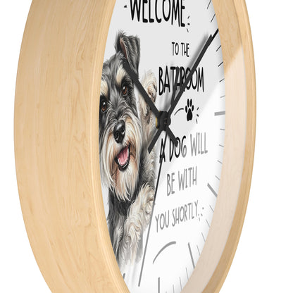 A round wall clock with a light natural wood frame and a white face featuring a detailed illustration of a gray and white dog. The clock face includes black text that reads 'WELCOME TO THE BATROOM A DOG WILL BE WITH YOU SHORTLY' along with a small paw print.