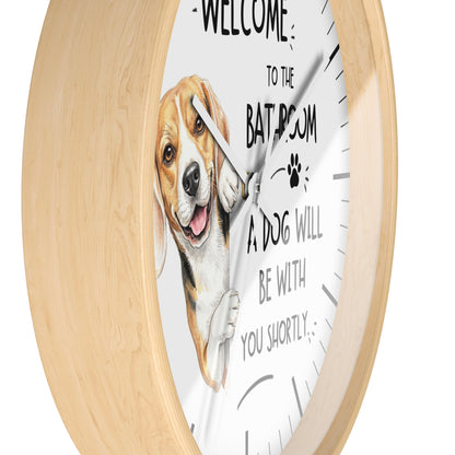 A round wall clock with a light natural wood frame and a white face. The clock face features a painted beagle dog illustration and the humorous text 'WELCOME TO THE BATHROOM A DOG WILL BE WITH YOU SHORTLY'.