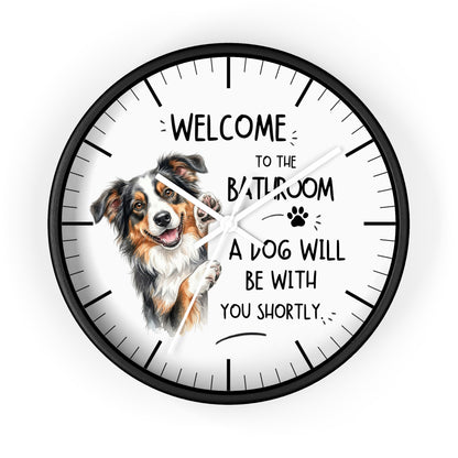 A round wall clock with a black frame and white face, featuring a watercolor illustration of a brown, white, and black dog on the left side. The clock face has black text that reads 'WELCOME TO THE BATHROOM A DOG WILL BE WITH YOU SHORTLY' along with a small paw print.