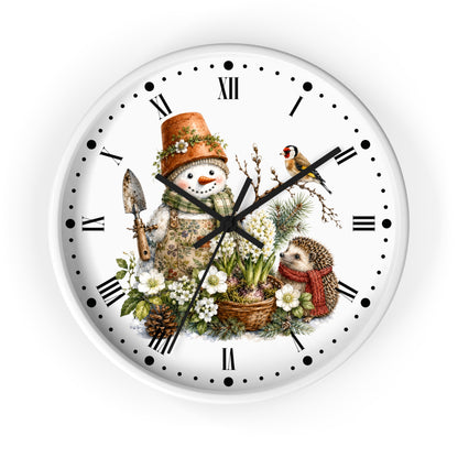 A round white wall clock with black Roman numerals and hands, featuring a whimsical winter garden illustration on the face. The design includes a snowman in a gardening hat with a trowel, a small bird, a hedgehog in a red scarf, flowers, pinecones, and greenery.