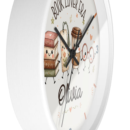 A round white wall clock with a cute book-themed illustration. The clock face features cartoon books, a coffee cup, and two glasses of juice with faces, along with the text 'BOOK LOVER ERA', the name 'Olivia', and decorative doodles in pink and gold.