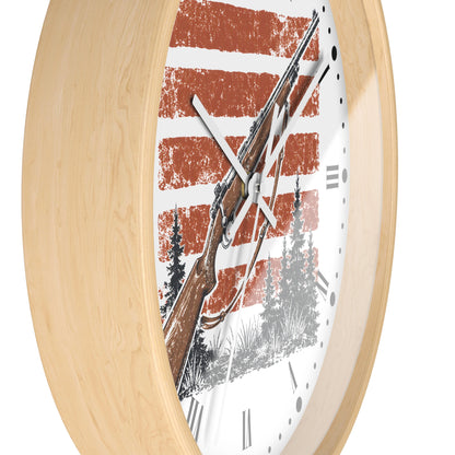 A close-up of a round wall clock with a light natural wood frame and a white face featuring a rustic red-and-white striped background, a brown rifle illustration, and black tree silhouettes. Roman numerals mark the hours.