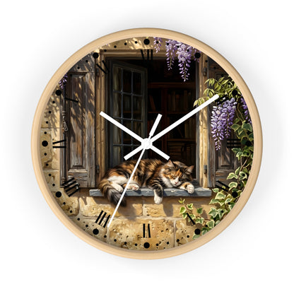 Round wall clock with a light wood frame featuring an illustrated scene of a fluffy orange tabby cat resting on a stone window sill, with purple wisteria flowers, green ivy, and dark wooden shutters. White clock hands and black Roman numerals complete the design.