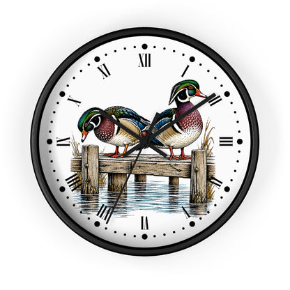 A round wall clock with a black frame and a white face featuring an illustration of two wood ducks on a wooden dock over water, with Roman numerals and black hands.
