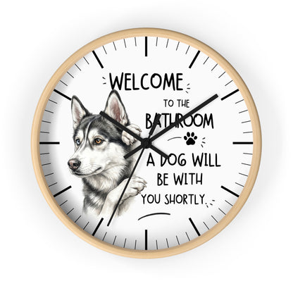 A round wall clock with a light wood frame and a white face, featuring a detailed illustration of a husky dog and the humorous text 'WELCOME TO THE BATHROOM A DOG WILL BE WITH YOU SHORTLY.'.