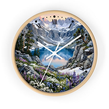 A round wall clock with a light natural wood frame and a scenic mountain landscape printed on the face. The artwork shows snow-capped peaks, a blue lake, rocky cliffs, evergreen trees, and wildflowers, with black Roman numeral hour markers and white clock hands.
